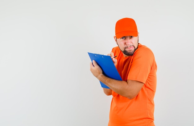 Delivery man in orange t-shirt, cap taking notes on clipboard and looking weird, front view.
