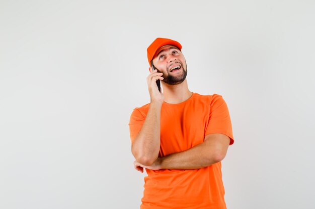 Delivery man in orange t-shirt, cap talking on mobile phone and looking cheerful , front view.