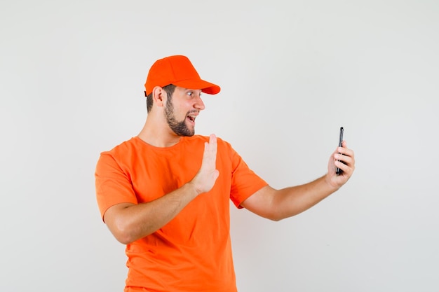 オレンジ色のTシャツ、ビデオ通話で手を振って陽気に見えるキャップ、正面図の配達人。