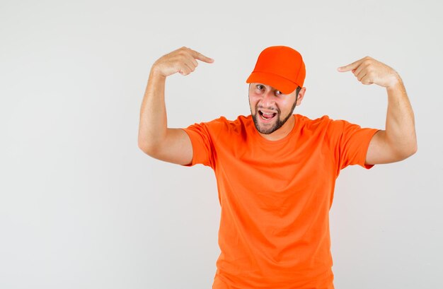 Delivery man in orange t-shirt pointing fingers at his cap and looking confident , front view.