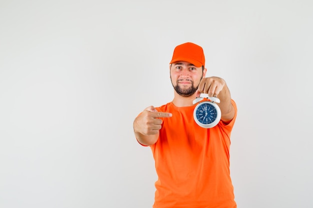 Delivery man pointing at alarm clock in orange t-shirt, cap and looking confident , front view.