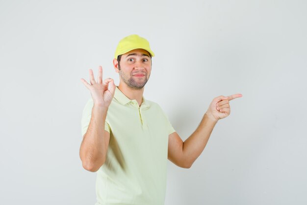 Delivery man pointing aside, showing ok sign in yellow uniform and looking confident. front view.