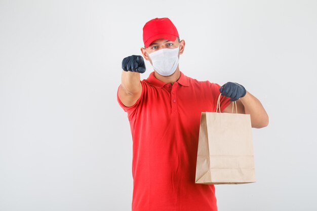 Delivery man pointing finger at camera with paper bag in red uniform, medical mask, gloves