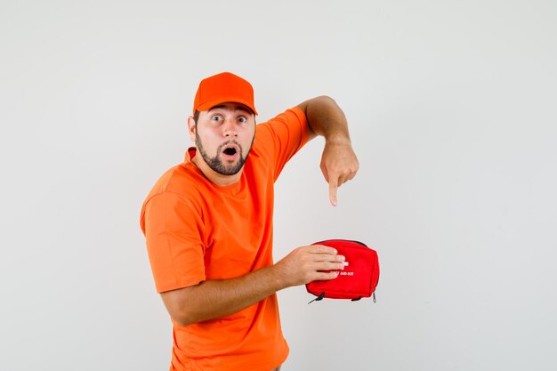 Delivery man pointing at inside of first aid kit in orange t-shirt, cap and looking anxious , front view.