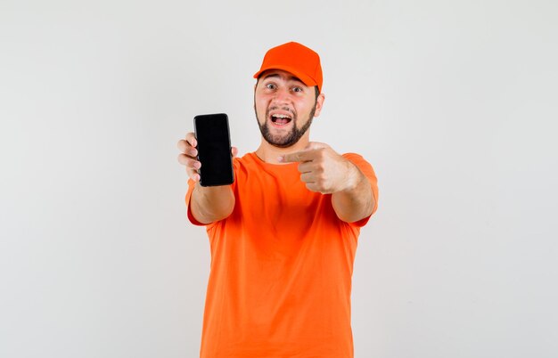 Delivery man pointing at mobile phone in orange t-shirt, cap and looking cheerful. front view.