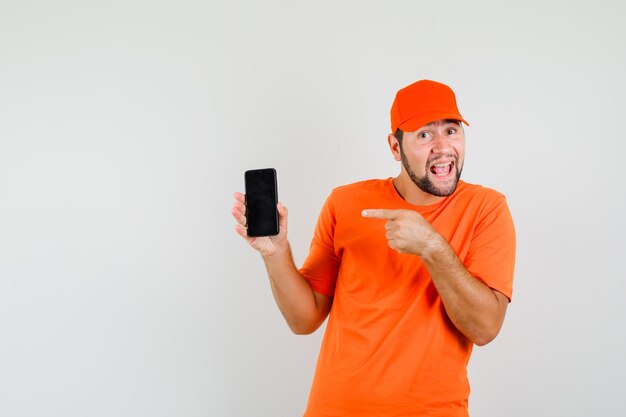 Delivery man pointing at mobile phone in orange t-shirt, cap and looking optimistic , front view.