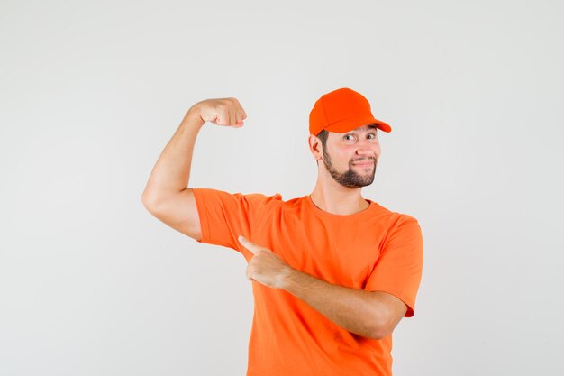 Доставщик, указывая на мышцы руки в оранжевой футболке, кепке и уверенно глядя, вид спереди.