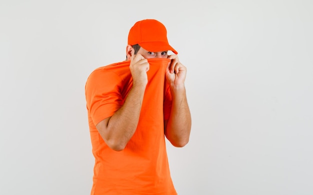 Delivery man pulling collar on face in orange t-shirt, cap and looking scared , front view.