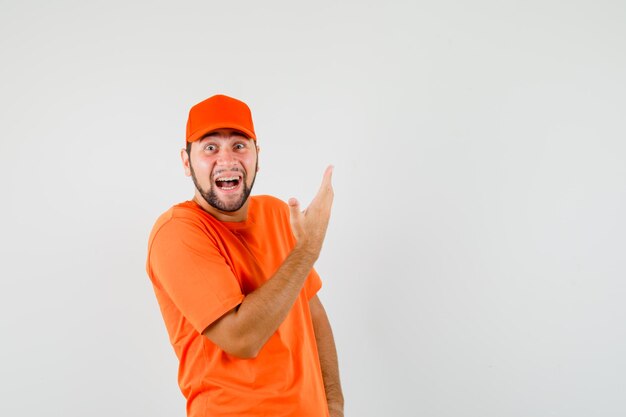 Delivery man raising arm in ecstatic gesture in orange t-shirt, cap and looking happy , front view.