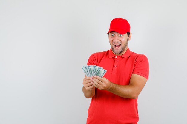 Delivery man in red t-shirt, cap holding dollar banknotes and looking happy