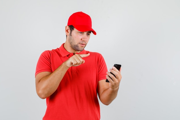 Delivery man in red t-shirt, cap looking at smartphone with finger sign