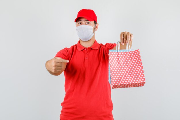 Доставщик в красной футболке, кепке, маске, указывая пальцем на бумажные пакеты