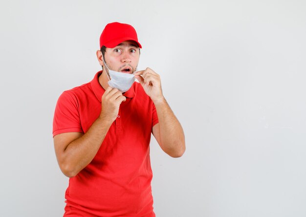 Delivery man in red t-shirt, cap taking off mask and looking surprised