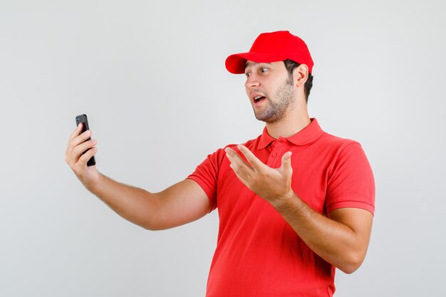 Delivery man in red t-shirt, cap talking on videocall and looking emotional