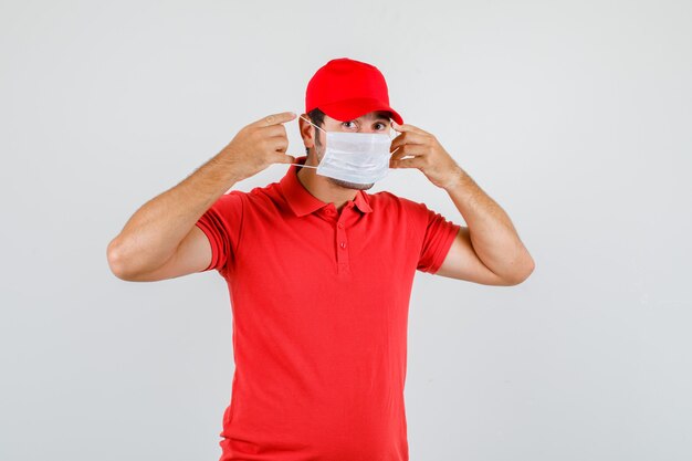 Delivery man in red t-shirt, cap wearing medical mask and looking careful