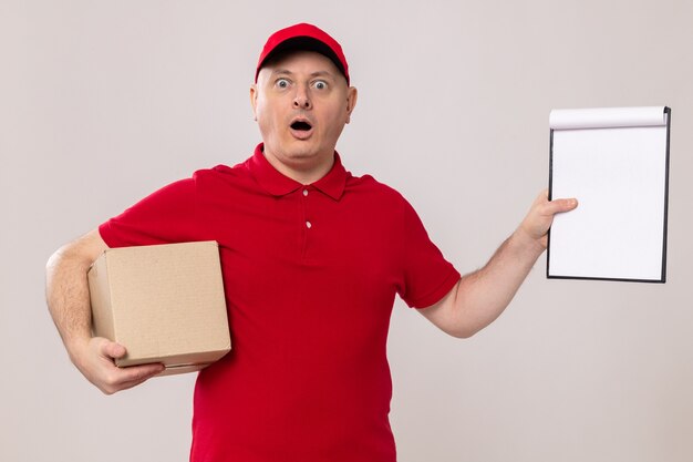 Delivery man in red uniform and cap holding cardboard box and clipboard with blank pages looking amazed and surprised