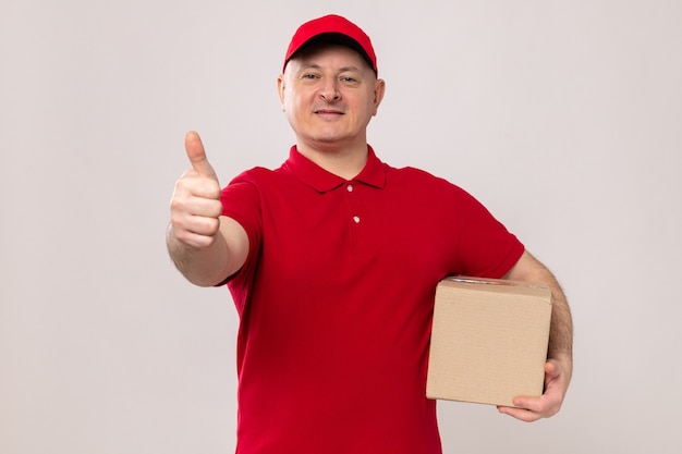 Доставщик в красной форме и кепке держит картонную коробку, глядя в камеру, уверенно улыбаясь, показывая большие пальцы руки вверх, стоя на белом фоне