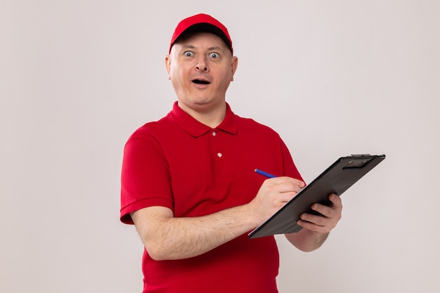 Delivery man in red uniform and cap holding clipboard and pen making notes looking at camera happy and excited standing over white background