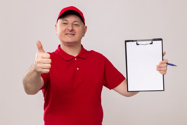 Delivery man in red uniform and cap holding clipboard with blank pages looking at camera smiling confident showing thumbs up standing over white background