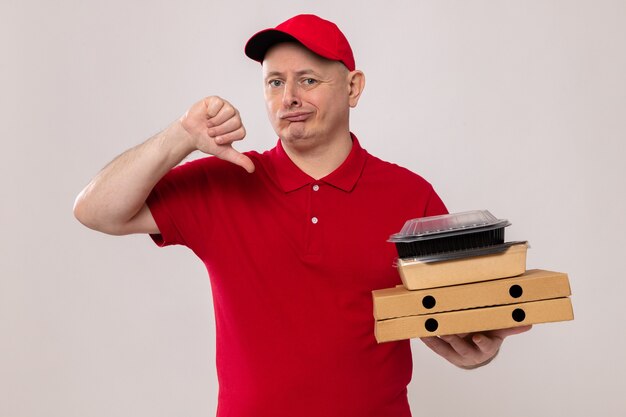 Delivery man in red uniform and cap holding pizza boxes and food packages looking displeased showing thumbs down