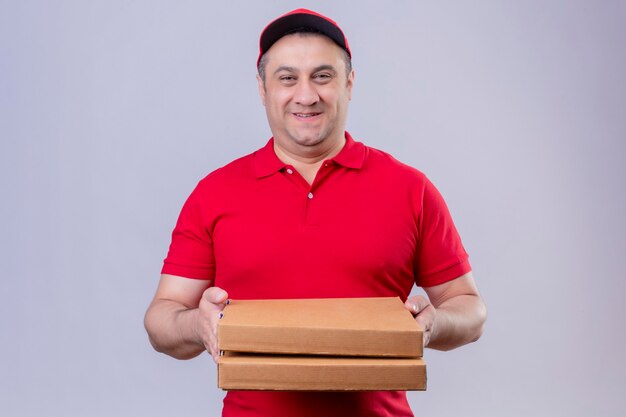 Доставщик в красной форме и кепке держит коробки для пиццы, выглядящие позитивно и счастливые, улыбающиеся, дружелюбные, стоя над изолированным белым пространством
