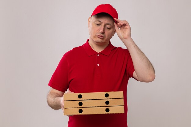 Delivery man in red uniform and cap holding pizza boxes looking at them puzzled standing over white background