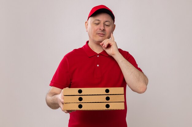 Delivery man in red uniform and cap holding pizza boxes looking at them with pensive expression smiling thinking positive