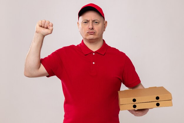 Delivery man in red uniform and cap holding pizza boxes looking with serious confident expression raising fist like a winner