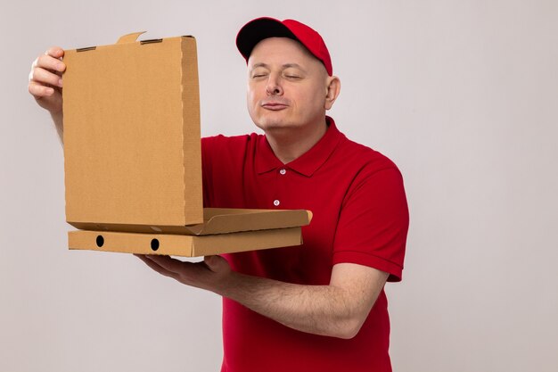 Delivery man in red uniform and cap holding pizza boxes opening one of them inhaling pleasant aroma