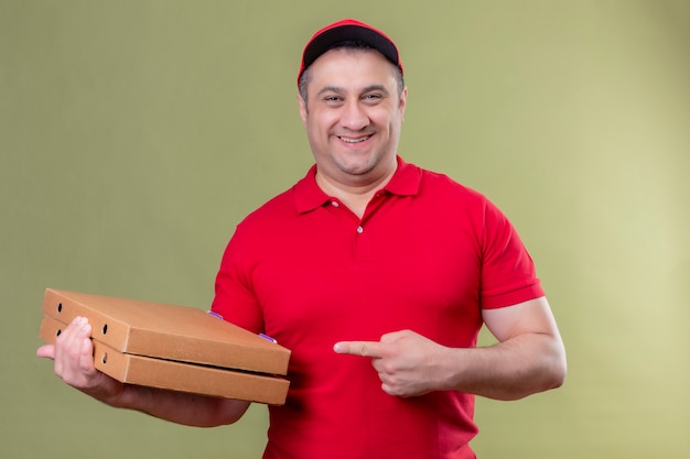 Fattorino in uniforme rossa e cappuccio che tiene le scatole per pizza che punta con il dito indice a loro sorridendo fiducioso con la faccia felice in piedi sopra lo spazio verde