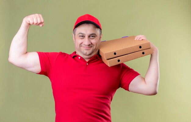 Delivery man in red uniform and cap holding pizza boxes showing biceps on his hand smiling confident winner concept standing over green space