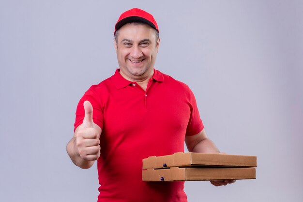 Delivery man in red uniform and cap holding pizza boxes  with happy face showing thumbs up standing over isolated white space