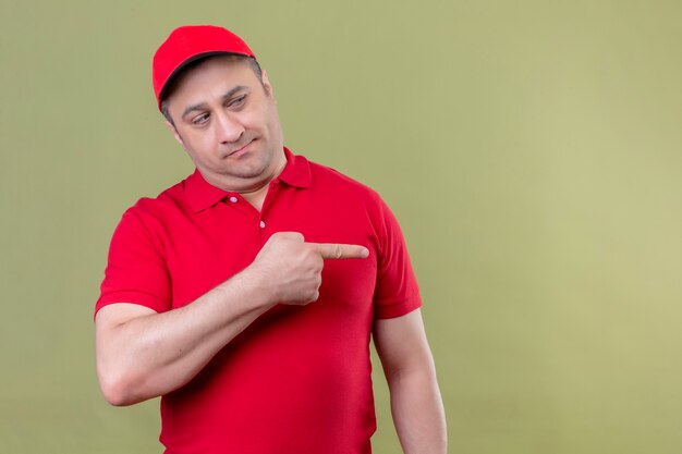빨간 제복을 입은 배달 남자와 격리 된 녹색 공간 위에 서있는 측면을 검지 손가락으로 가리키는 회의적인 표정으로 옆으로 보이는 모자