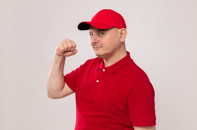 Delivery man in red uniform and cap looking at camera with confident expression raising fist like a winner standing over white background