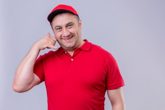 Delivery man in red uniform and cap smiling cheerfully making call me gesture standing on white