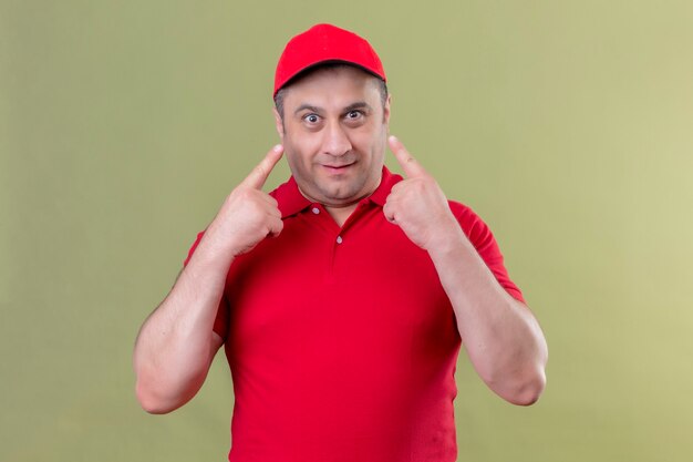 Delivery man in red uniform and cap  smiling pointing his eyes standing over green space