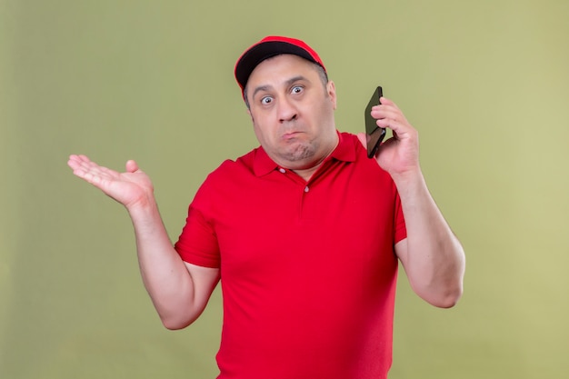 Free photo delivery man in red uniform and cap standing with smartphone shrugging shoulders, spreading hands not understanding what happened clueless and confused expression standing