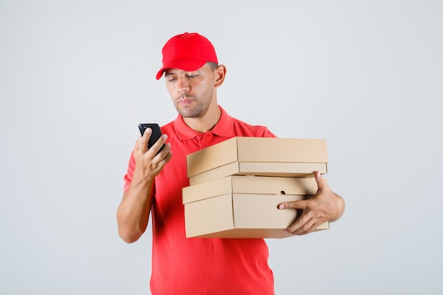 Delivery man in red uniform holding cardboard boxes while using smartphone