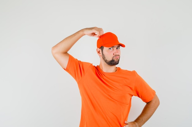 Delivery man scratching head in orange t-shirt, cap and looking pensive. front view.