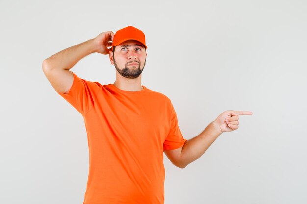 Delivery man scratching head, pointing to the side in orange t-shirt, cap and looking pensive. front view.