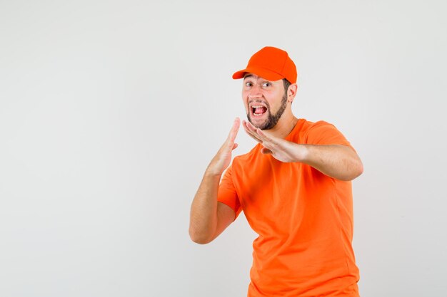 Delivery man showing karate chop gesture in orange t-shirt, cap and looking powerful. front view.