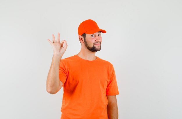 Delivery man showing ok sign in orange t-shirt, cap and looking cheery. front view.