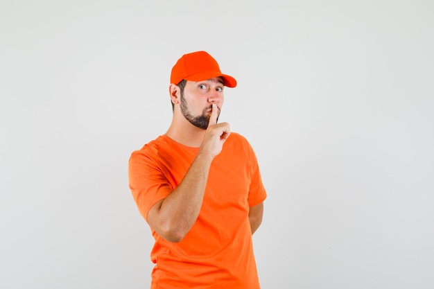 Delivery man showing silence gesture in orange t-shirt, cap and looking careful. front view.