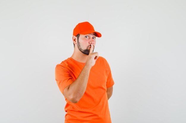 Доставщик показывает жест молчания в оранжевой футболке, кепке и внимательно смотрит. передний план.