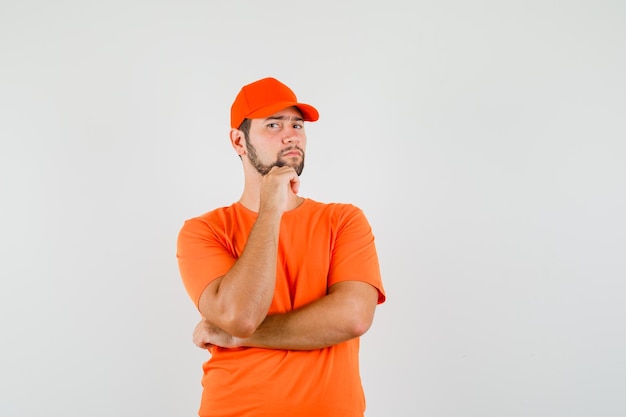 Delivery man standing in thinking pose in orange t-shirt, cap and looking sensible , front view.