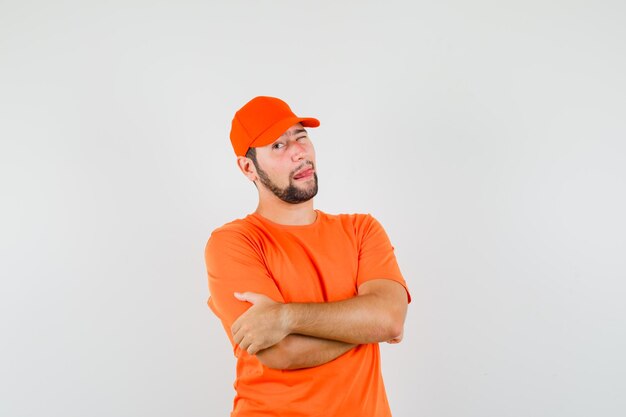 Delivery man standing with crossed arms, winking eye, sticking out tongue in orange t-shirt, cap front view.