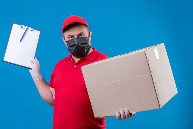Delivery man wearing red uniform and cap in facial protective mask holding large cardboard box and clipboard  with serious face standing over blue space
