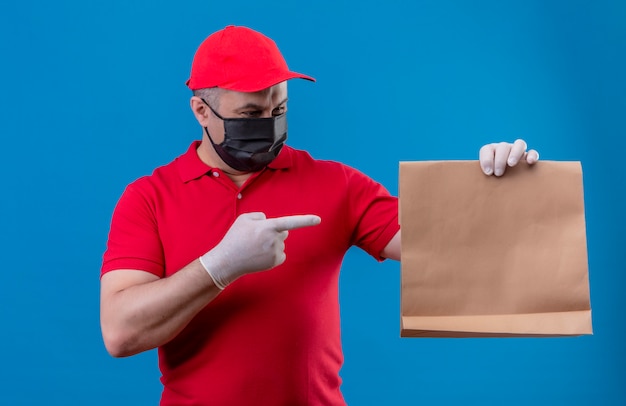 Delivery man wearing red uniform and cap in facial protective mask holding paper package pointing with index finger to it smiling over isolated blue wall