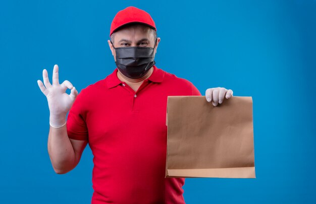 Доставка человек в красной форме и кепке в защитной маске для лица держит бумажный пакет положительным и счастливым делать хорошо подписать через синюю стену