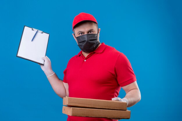 Доставщик в красной форме и кепке в защитной маске для лица держит коробки для пиццы и буфер обмена с серьезным лицом, стоящим над синим пространством